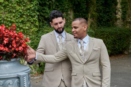 Foto de Un par de hombres de la comunidad lgbt pasear por el jardín después de su boda, ambos están felizmente mirando algunas flores. - Imagen libre de derechos