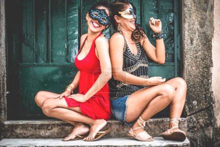 Photo for Portrait of two beautiful and happy girls sitting against a green colored door. City girls far from the capital. They handle a carnival mask. - Royalty Free Image