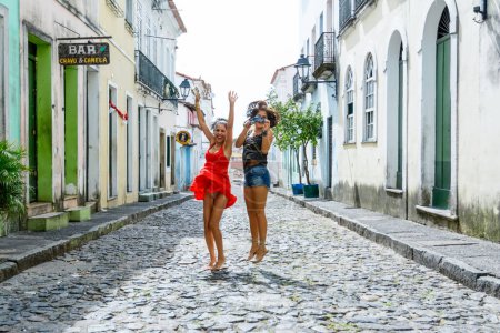 Photo for Portrait of two beautiful and happy girls jumping on the cobblestone street. Pelourinho streets in Salvador, Brazil. - Royalty Free Image