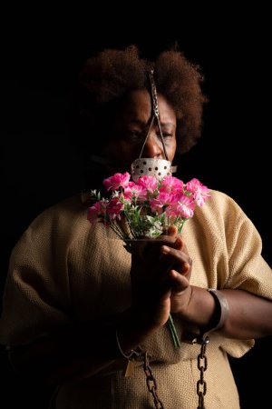 Photo for Portrait of a black woman with chains on her hands and an iron mask on her face holding a bunch of flowers. Slave Anastacia. Slavery in Brazil. - Royalty Free Image