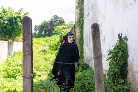 Photo for Santo Amaro, Bahia, Brazil - July 24, 2022: A person walks among the plants dressed in black with a mask of terror during a cultural demonstration in Acupe in the city of Santo Amaro, Bahia. - Royalty Free Image