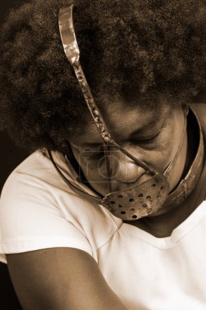 Photo for Black woman in chains with an iron mask on her face, representing the slave Anastacia. Pain and suffering, torture. Against black background. - Royalty Free Image