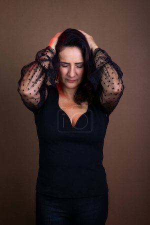 Photo for Coaching woman in black clothing with hands on the back of her head in a sign of concern. Isolated on brown background. - Royalty Free Image