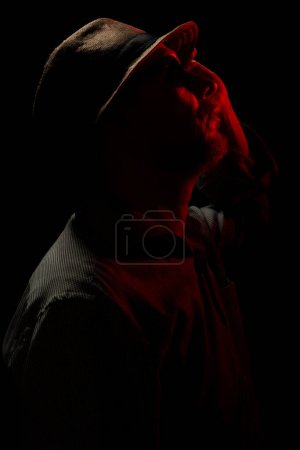 Foto de Retrato discreto del hombre con sombrero y gafas de sol en la oscuridad con luz roja en la cara. Arte habitación oscura. - Imagen libre de derechos