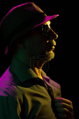 Foto de Retrato de un hombre barbudo serio con sombrero y gafas de sol. Retrato en estudio oscuro. - Imagen libre de derechos