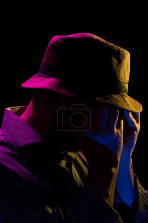 Foto de Retrato de un hombre serio y misterioso con barba con sombrero y gafas de sol haciendo gestos con las manos. Retrato en estudio con luces de colores y fondo oscuro. - Imagen libre de derechos