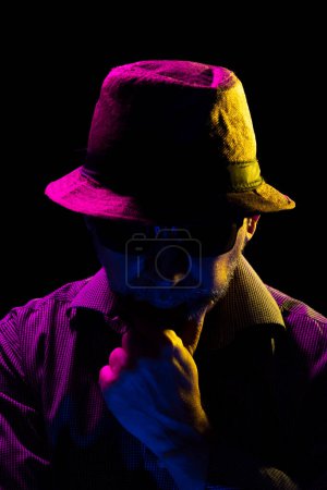 Foto de Retrato de un hombre serio y misterioso con una barba con sombrero y gafas de sol tocándose la cara. Retrato en estudio con luces de colores y fondo oscuro. - Imagen libre de derechos