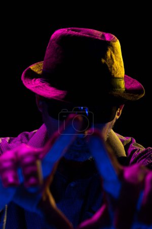 Foto de Retrato de un hombre serio y misterioso con barba con sombrero y gafas de sol haciendo gestos con las manos. Retrato en estudio con luces de colores y fondo oscuro. - Imagen libre de derechos