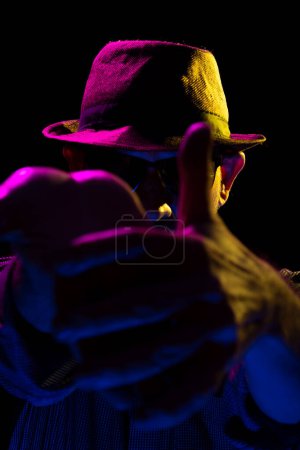Foto de Misterioso hombre barbudo serio con sombrero y gafas de sol haciendo gestos con las manos. Retrato en estudio con luces de colores y fondo oscuro. - Imagen libre de derechos