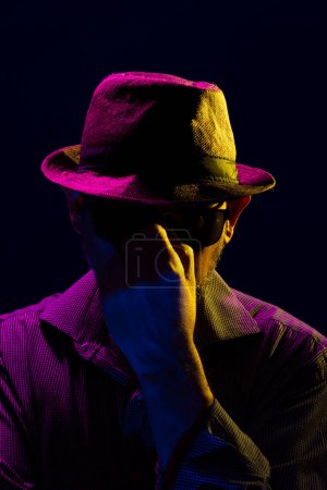 Foto de Retrato de un hombre serio y misterioso con una barba con sombrero y gafas de sol tocándose la cara. Retrato en estudio con luces de colores y fondo oscuro. - Imagen libre de derechos
