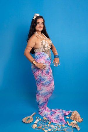 Beautiful woman, with black hair, wearing a tiara and bracelets made of sea shells, against a blue background, dressed as a mermaid next to several shells, a mirror and pearl necklaces. She is a devotee of iemanja.