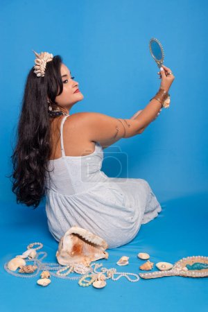 Schöne schwarzhaarige Frau in weißer Kleidung, die vor blauem Hintergrund neben mehreren Muscheln sitzt, ihren Rücken hält und in einen Spiegel blickt. Sie ist eine Anhängerin von iemanja.