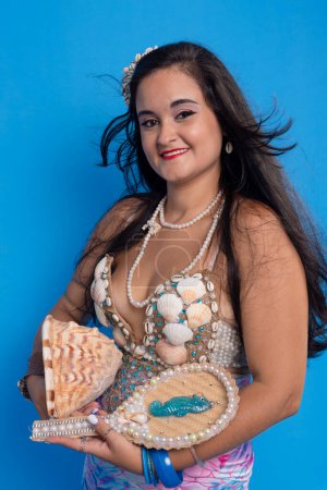 Beautiful woman, wearing a tiara and accessories, against a blue background, holding a mirror and a sea shell. Yemanja worshiper.