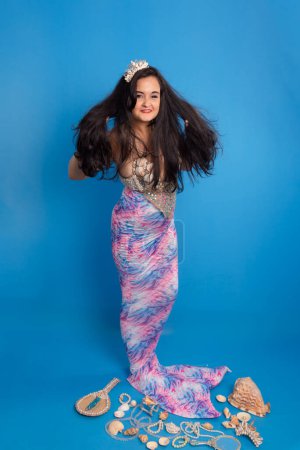 Hermosa mujer, llevando una tiara y pulseras hechas de conchas marinas, sobre un fondo azul, vestida de sirena junto a varias conchas, un espejo y collares de perlas tocando su pelo negro. Ella es una devota de iemanja.