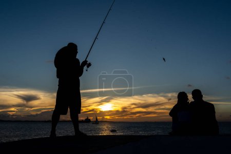 Foto de Salvador, Bahia, Brasil - 13 de abril de 2019: Pescador es visto en silueta durante la puesta de sol en Ponta do Humaita en la ciudad de Salvador, Bahia. - Imagen libre de derechos