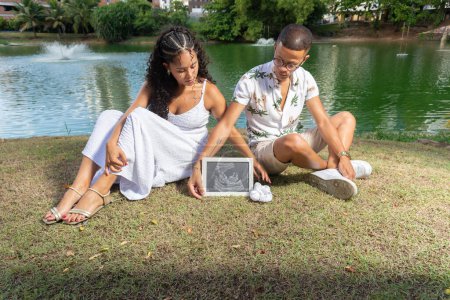 Téléchargez les photos : Couple assis au bord d'un lac, en vêtements blancs, montrant un test de grossesse encadrée. Elle est enceinte. Genre révélé. En arrière-plan une lagune verte. - en image libre de droit
