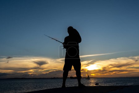 Foto de Salvador, Bahia, Brasil - 13 de abril de 2019: Una persona es vista, en silueta, pescando al atardecer en Ponta do Humaita en la ciudad de Salvador, Bahia. - Imagen libre de derechos