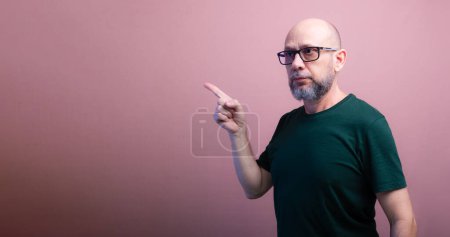 Bearded bald man with prescription glasses pointing with his finger to the side. Isolated on salmon colored background.