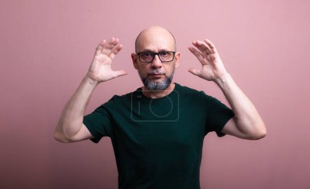 Portrait of bald, bearded man with glasses with both hands next to his head looking at the camera. Isolated on salmon colored background.