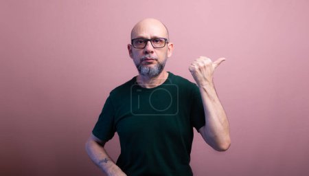 Portrait of bearded bald man wearing prescription glasses pointing to the side with his thumb. Isolated on salmon colored background.
