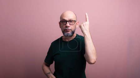 Bearded bald man with prescription glasses pointing up with his finger. Isolated on salmon colored background.