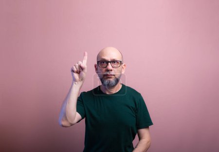 Bearded bald man with prescription glasses pointing up with his finger. Isolated on salmon colored background.