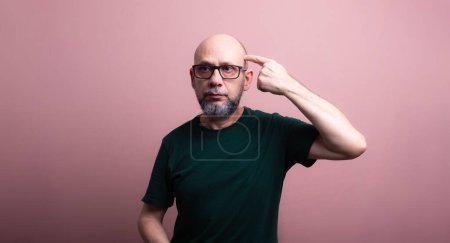 Bearded bald man wearing prescription glasses pointing to his head with his finger. Isolated on salmon colored background.