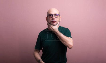 Portrait of a bald, bearded man wearing prescription glasses squeezing his neck with one hand. Isolated on salmon colored background.