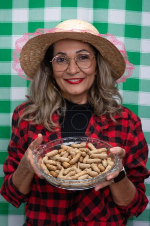 Woman in typical clothes wearing a straw hat holding a glass tray with peanuts inside. Food from the Sao Joao festival. Isolated on green and white checkered background.