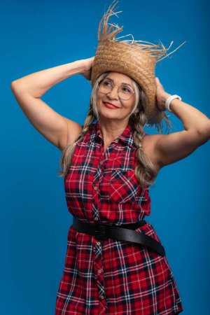 Beautiful woman, dressed for the Sao Joao festival, standing, holding a straw hat and dancing. Isolated on blue background.
