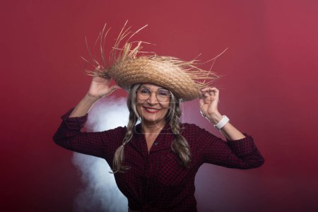 Beautiful woman with braided hair holding a straw hat in typical clothing for the Sao Joao festival, dancing and posing. Isolated on smoky red background.