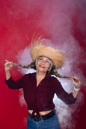 Femme portant un chapeau de paille avec des vêtements typiques pour le festival de Sao Joao dansant et tenant ses tresses de cheveux. Isolé sur fond rouge fumé.