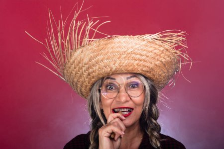 Face portrait of beautiful woman with braided hair wearing straw hat and eating boiled peanuts. Celebration of the feast of Saint John. Isolated on smoky red background.