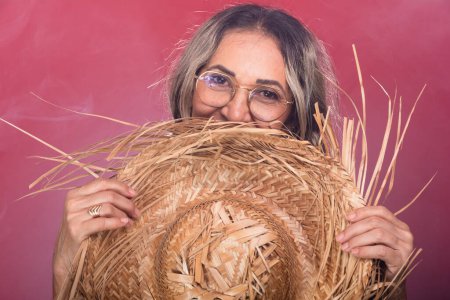 Close-up portrait of a beautiful woman with glasses and braided hair holding a straw hat in typical clothing for the Sao Joao festival. Isolated on smoky red background.