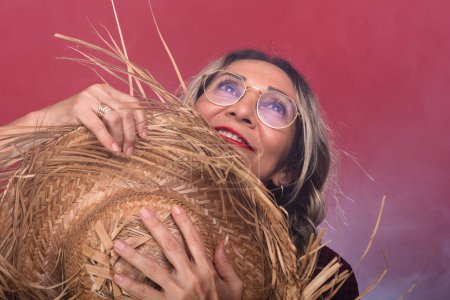 Close-up portrait of a beautiful woman with glasses and braided hair holding a straw hat in typical clothing for the Sao Joao festival. Isolated on smoky red background.