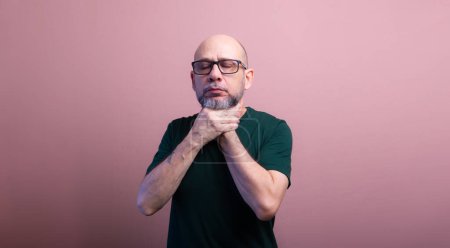 Bearded bald man with prescription glasses squeezing his neck with both hands. Isolated on salmon colored background.