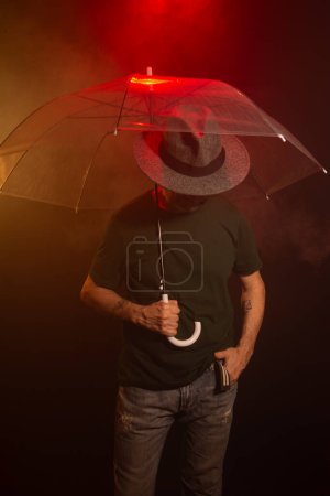 Geheimnisvoller Mann mit Hut und durchsichtigem Regenschirm. Studioporträt. Roter dunkler Hintergrund mit künstlichem Rauch.