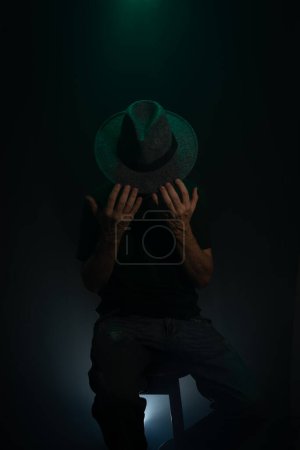 Mysterious, bearded man wearing a hat posing for the camera. Studio portrait. Dark green background with artificial smoke.