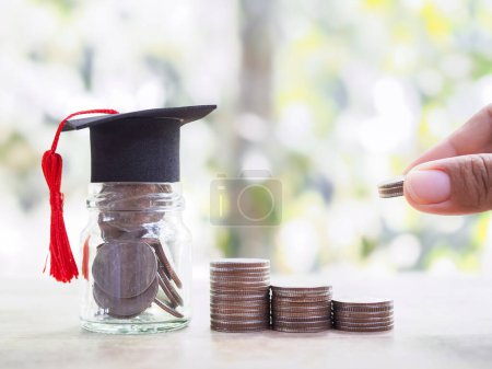 Glass bottle with graduation hat and hand putting coin into stack of coins. The concept of saving money for education, student loan, scholarship, tuition fees in the future