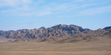 un large désert avec une vaste plaine plate s'étend, couverte d'une végétation clairsemée et sèche et de collines vallonnées, menant à une chaîne de montagnes accidentée avec la lumière du soleil créant un jeu d'ombres.