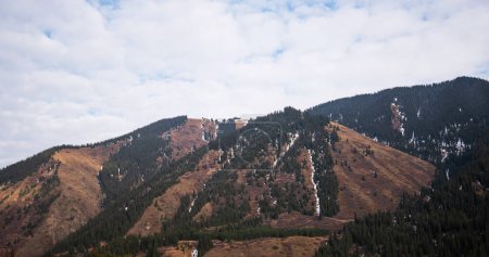 un paysage montagneux avec un mélange de forêts à feuilles persistantes et de zones herbeuses brunes, entrecoupées de plaques de neige sous un ciel nuageux.