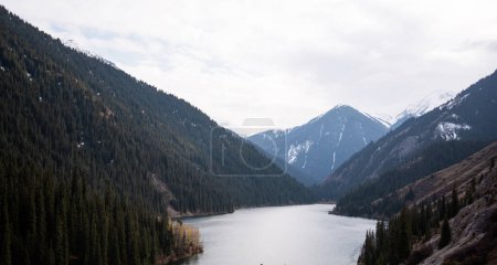 un lago de montaña situado en un valle, flanqueado por laderas boscosas y picos nevados bajo un cielo tenue y nublado, que transmite una sensación de aislamiento pacífico