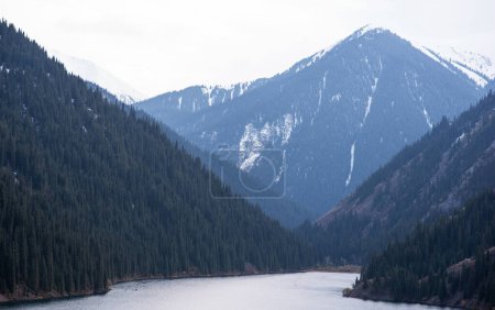 a mountain lake nestled in a valley, flanked by forested slopes and snowy peaks under a subdued, cloudy sky, conveying a sense of peaceful isolation