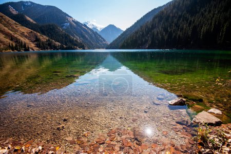 Les eaux cristallines d'un lac de montagne révèlent des pierres en dessous, conduisant à des collines boisées et à des sommets enneigés sous un soleil éclatant dans un ciel bleu profond.