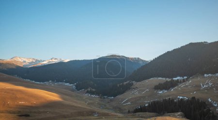 Dusk light illuminates a mountainous landscape, highlighting a winding valley with sparse snow and dense coniferous forests leading to distant peaks.