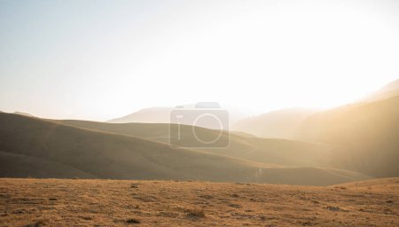 Soft sunlight filters over undulating grassy hills, casting a warm glow on the serene highland landscape with distant mountain silhouettes.