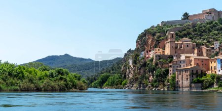a picturesque riverside village Miravet, Spain. Ancient buildings clinging to the steep hillside, the river flows calmly in the foreground, framed by the lush greenery of the surrounding landscape..