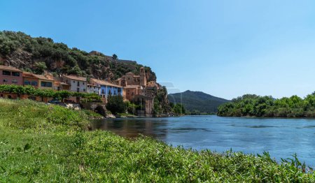 une rivière sereine qui coule devant un village perché, avec l'architecture du village qui se fond parfaitement dans le paysage naturel luxuriant sous un ciel bleu clair