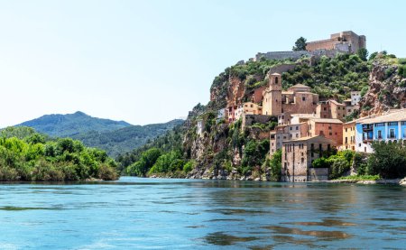 a picturesque riverside village Miravet, Spain. Ancient buildings clinging to the steep hillside, the river flows calmly in the foreground, framed by the lush greenery of the surrounding landscape.