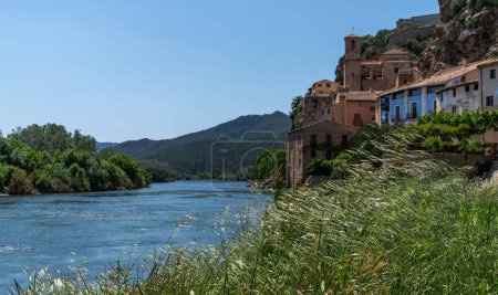 Ein ruhiger Fluss fließt sanft an einem historischen Dorf mit Terrakottadächern vorbei, eingebettet in einen Hintergrund üppiger Hügel unter einem klaren blauen Himmel, der die Essenz ländlicher Ruhe einfängt.